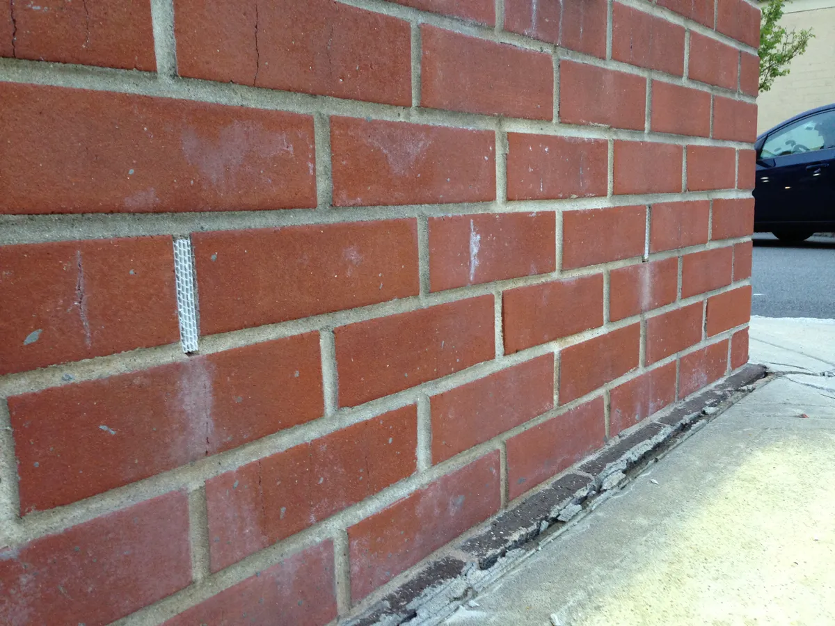 Weep holes fitted with white plastic grid inserts in the vertical mortar joints of a red brick wall, just above ground level